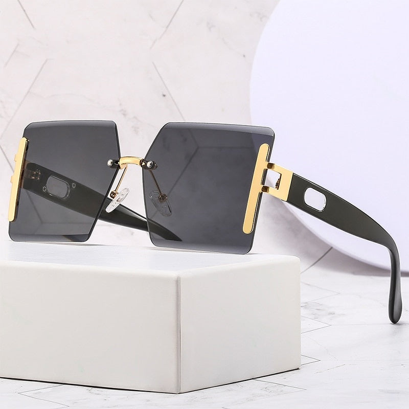 Sunglasses with black lenses and gold frames on a white box with a light gray geometric patterned background.
