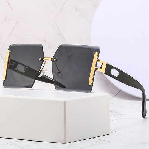 Sunglasses with black lenses and gold frames on a white box with a light gray geometric patterned background.